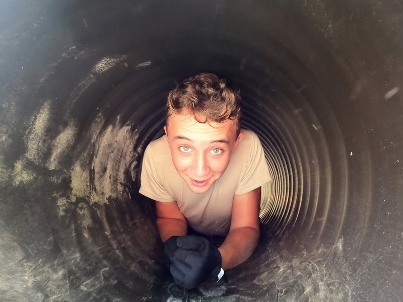 A Civil Air Patrol cadet grinning up at the camera while crawling through a drainage culvert during field training