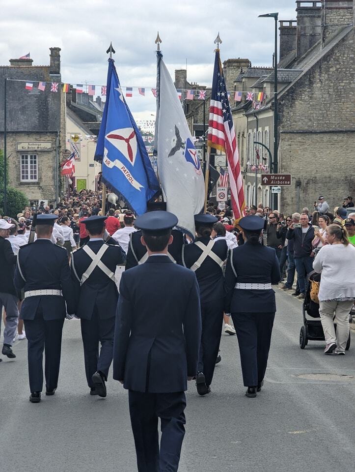 The squadron color guard carrying the American flag through a crowd-lined street during a commemorative parade in France